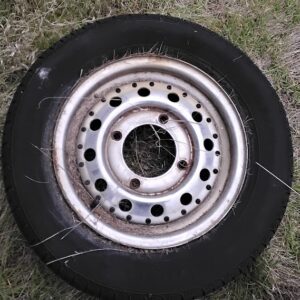 A vintage 4-lug Subaru wheel rim with a classic 4x140mm bolt pattern, mounted with a tire and lying flat on a patch of grass,