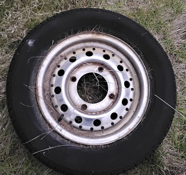 A vintage 4-lug Subaru wheel rim with a classic 4x140mm bolt pattern, mounted with a tire and lying flat on a patch of grass,